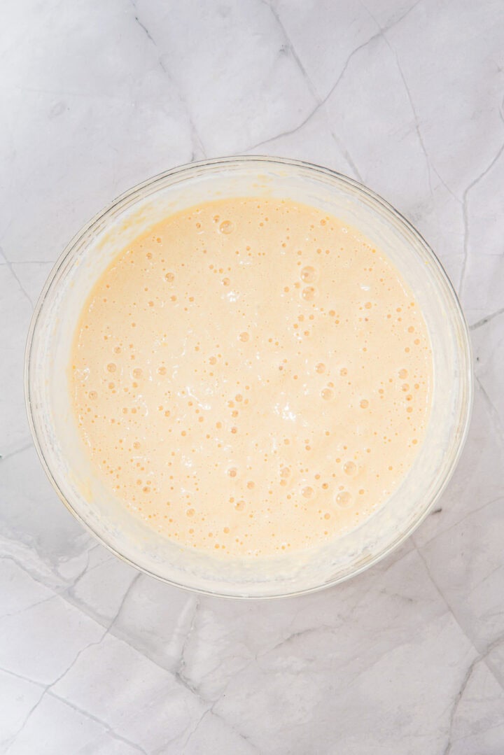 A glass bowl filled with smooth, bubbly pancake batter on a light marble surface.