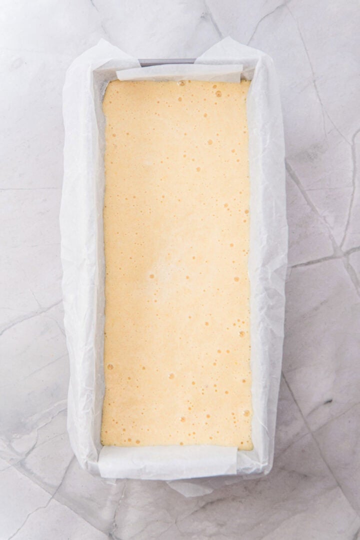 Rectangular baking pan lined with parchment paper, filled with pale yellow cake batter on a marble surface.