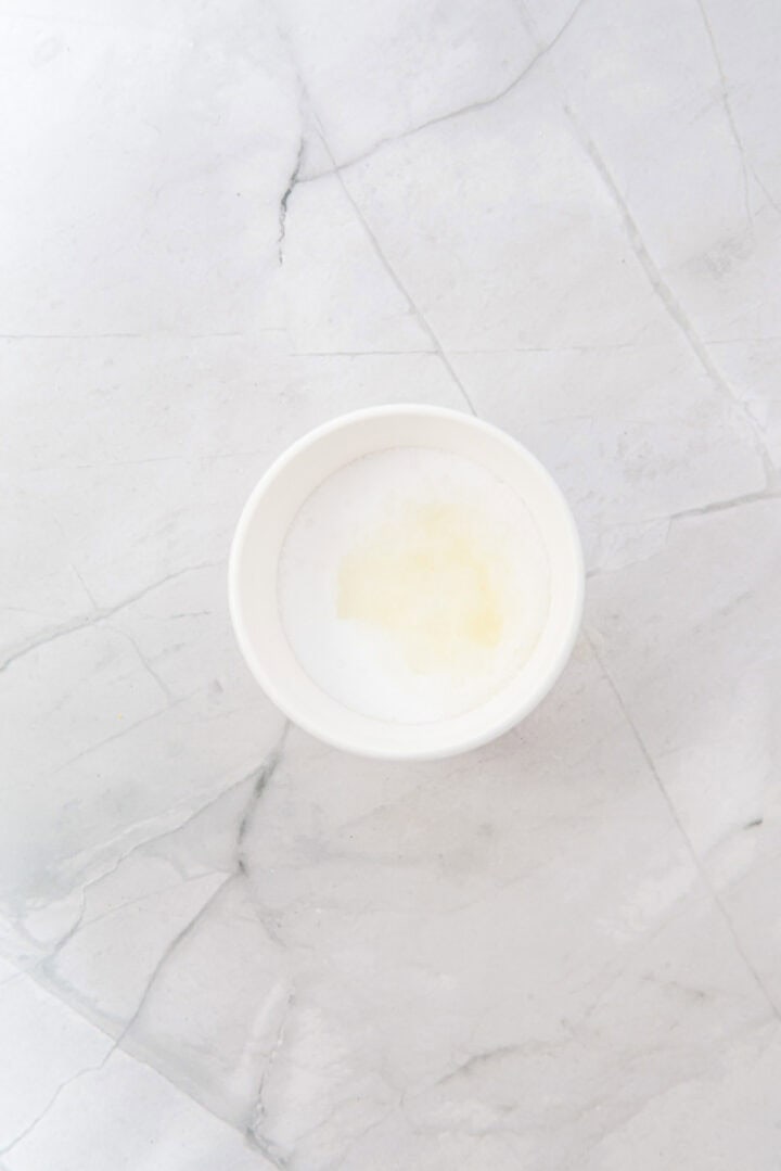A small white bowl with a light yellowish liquid on a white marble surface.