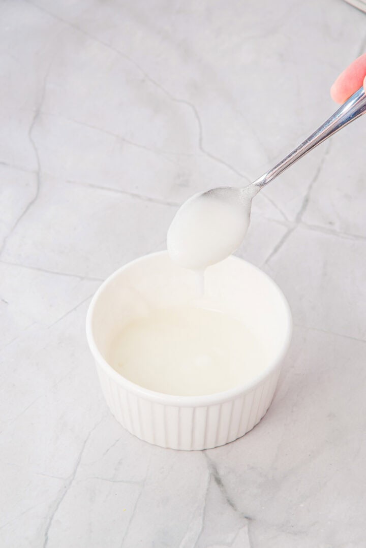 A hand holds a spoon dripping white icing into a small white ramekin on a marble surface.