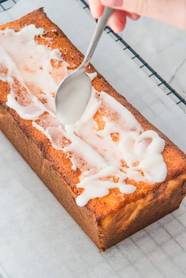 A hand spreads icing on a loaf cake with a spoon, on a wire rack lined with parchment paper.