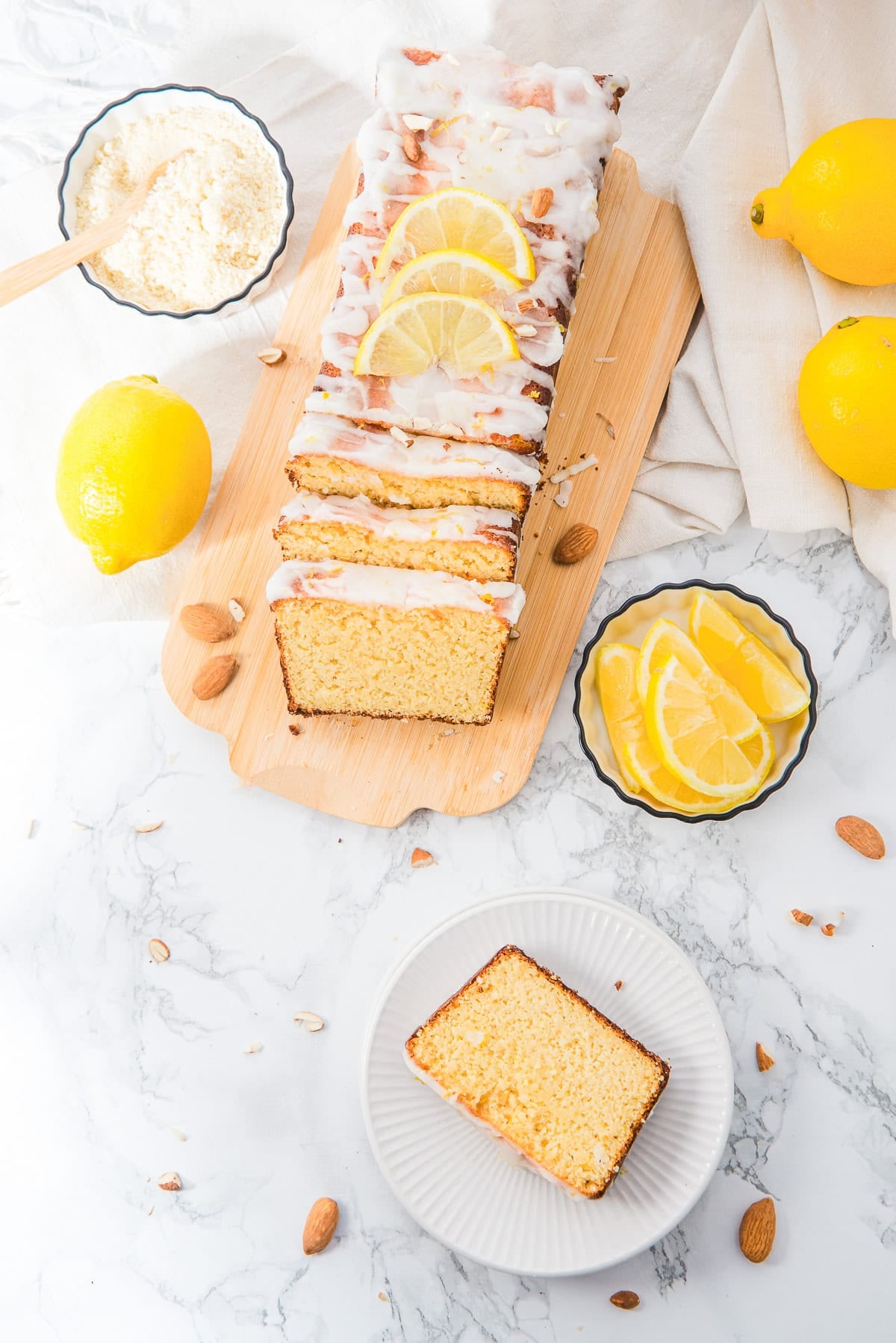 Lemon loaf cake with icing and lemon slices on top, surrounded by lemons, almonds, and a slice on a plate.