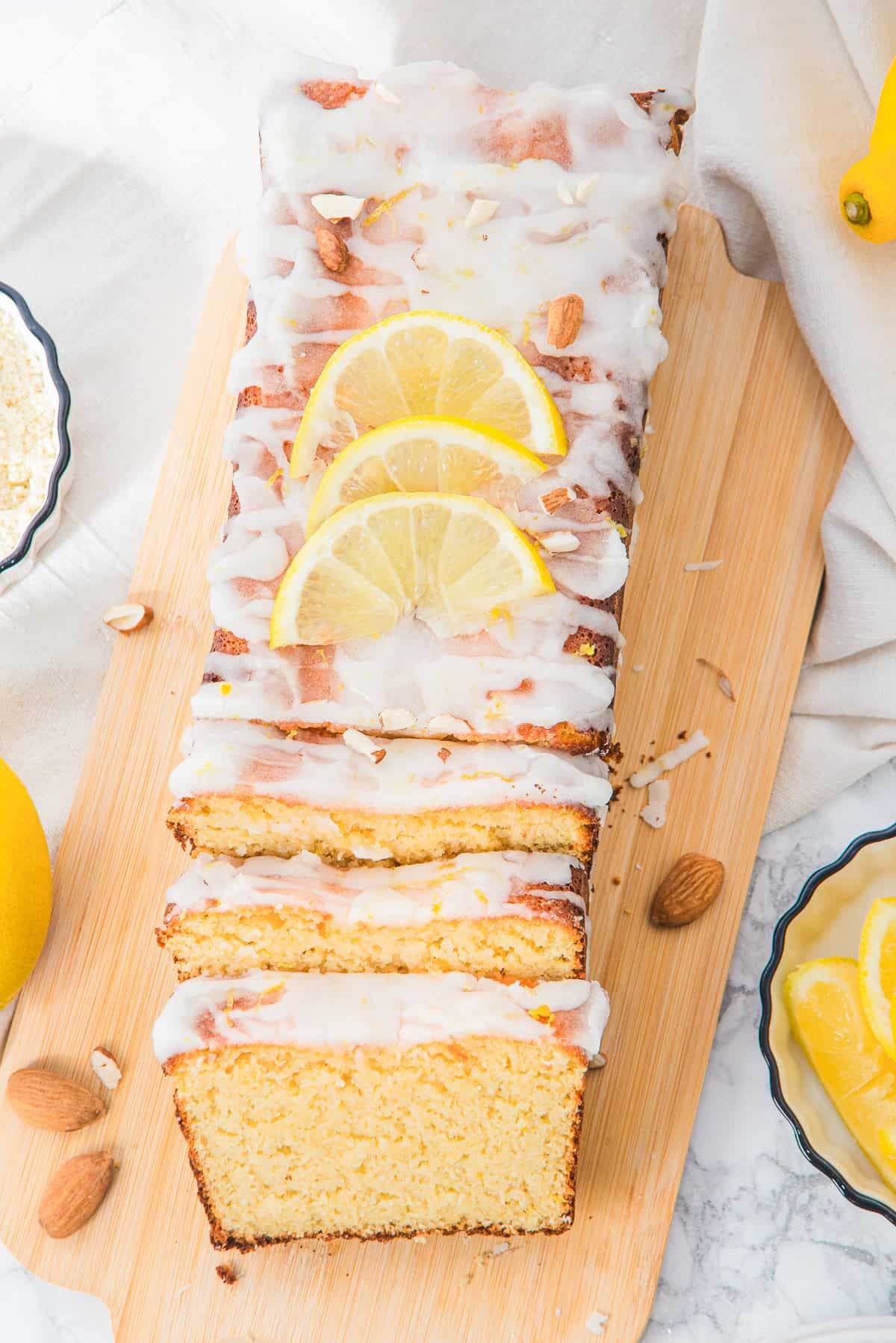 Lemon loaf cake with white icing, lemon slices, and almonds on a wooden board, partially sliced.