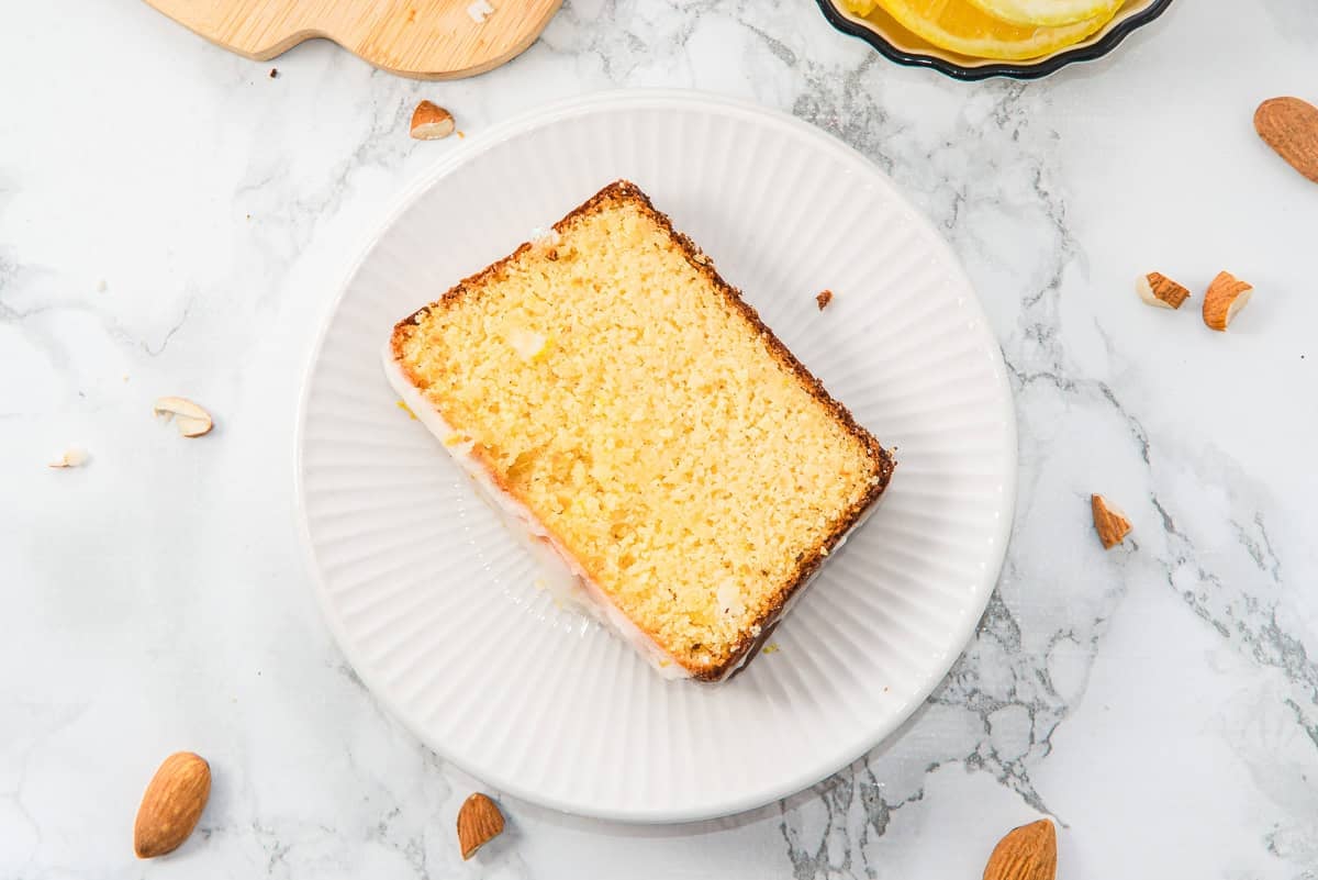A slice of Almond Flour Lemon Bread on a white plate, with almonds scattered on a marble surface.
