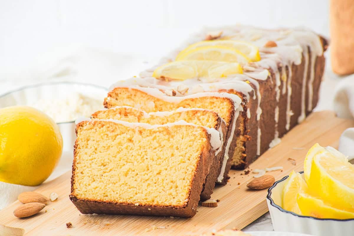 Sliced lemon loaf cake with icing and lemon slices on top, displayed on a wooden board with almonds and lemons nearby.