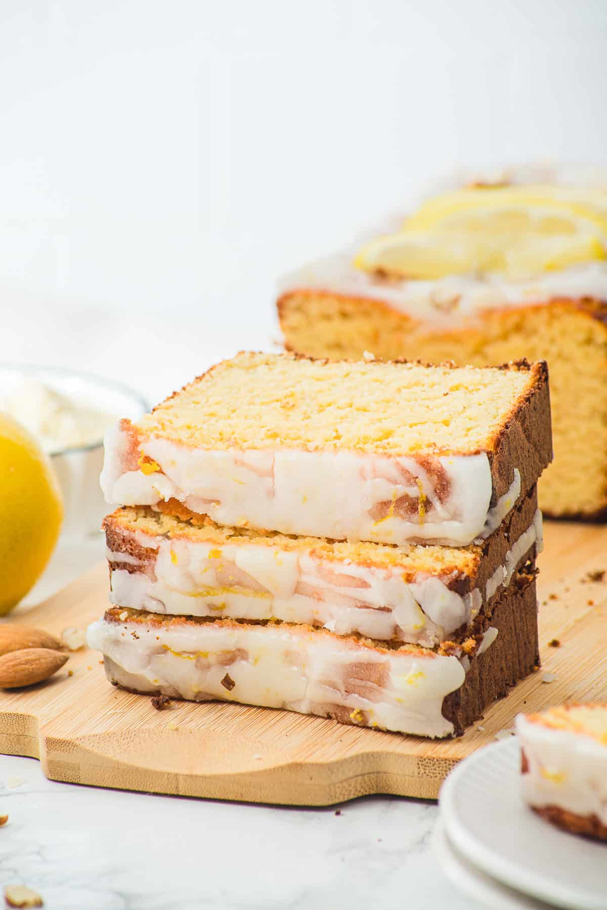 Three slices of glazed lemon loaf stacked on a wooden board with more loaf and a lemon in the background.