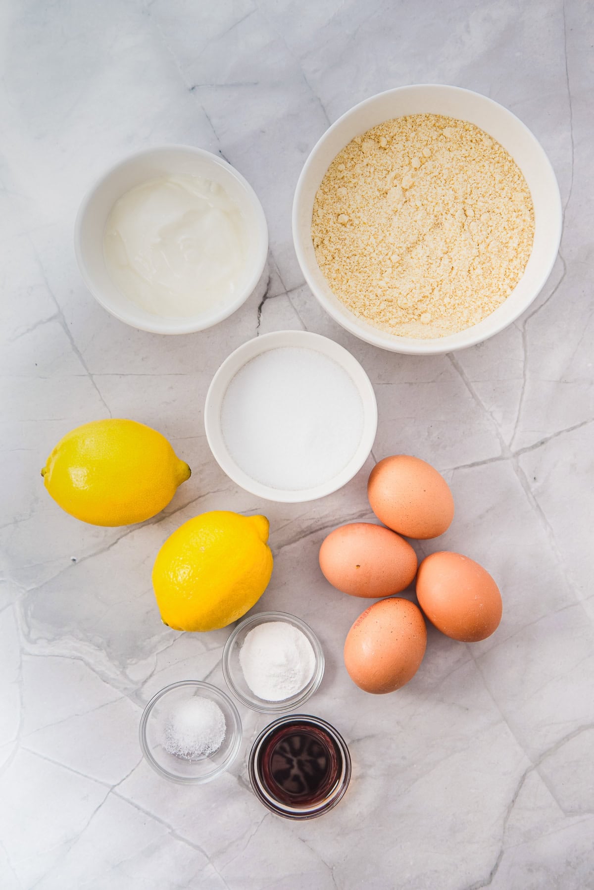 Top view of baking ingredients: cornmeal, eggs, lemons, sugar, yogurt, vanilla, salt, and baking powder on marble.