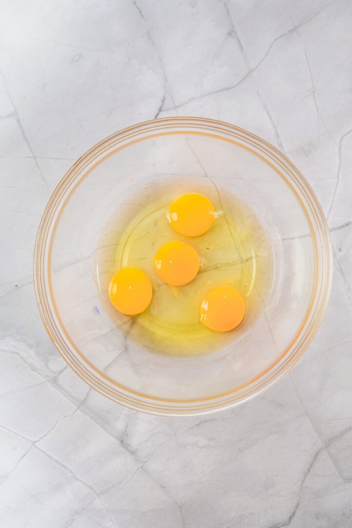 Four raw eggs cracked into a clear glass bowl on a light marble countertop.