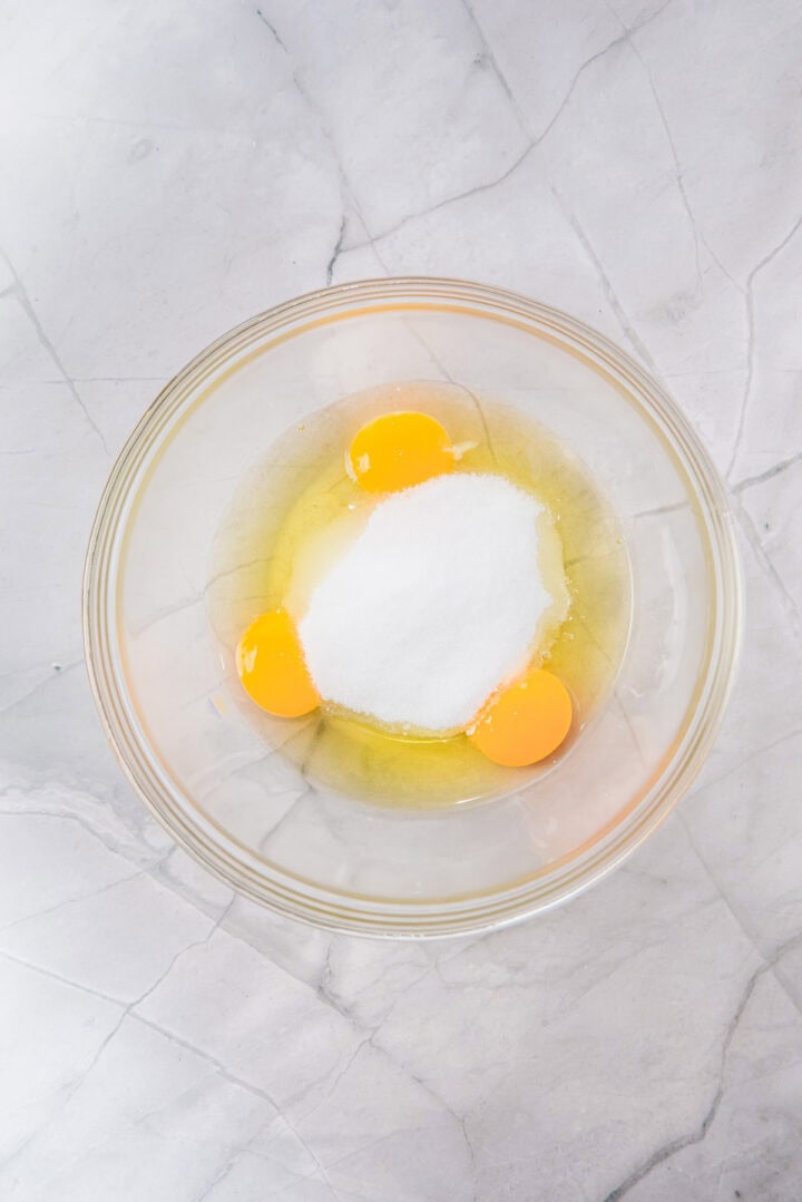 A glass bowl with three eggs and white sugar on a marble countertop.