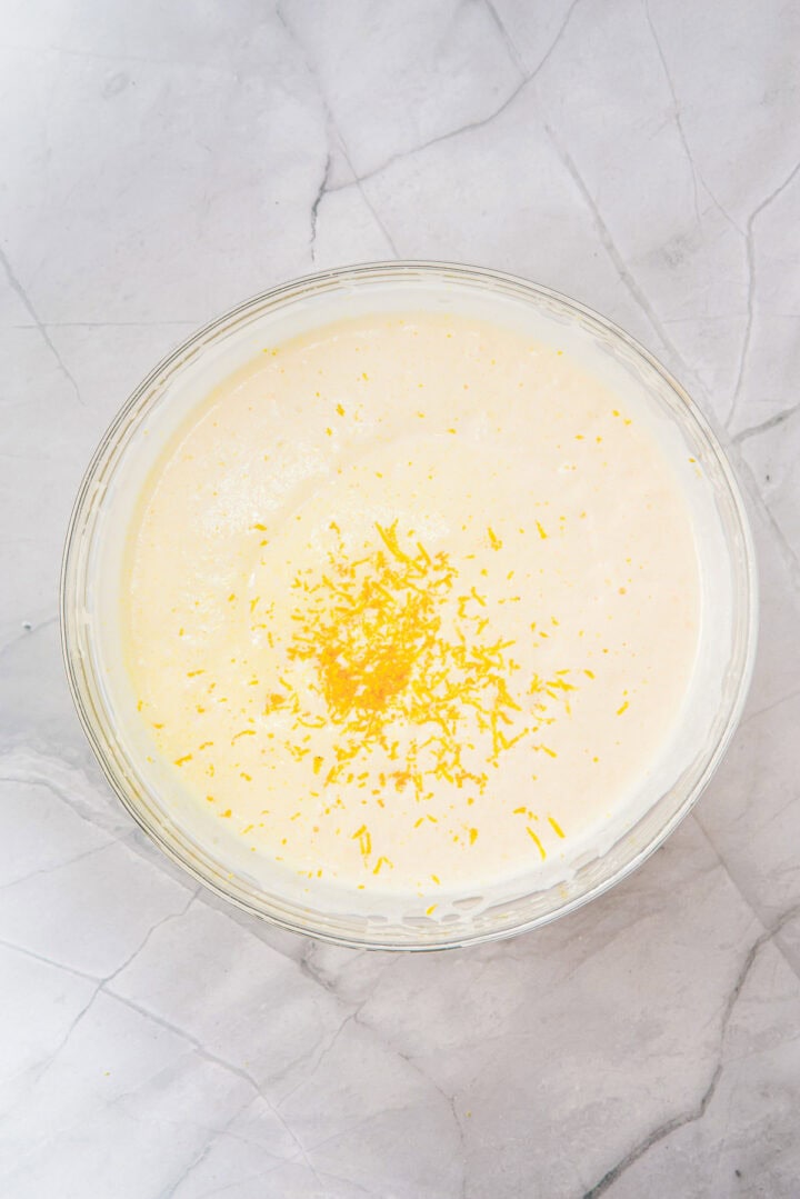 A glass bowl of creamy mixture topped with grated lemon zest on a marble surface.