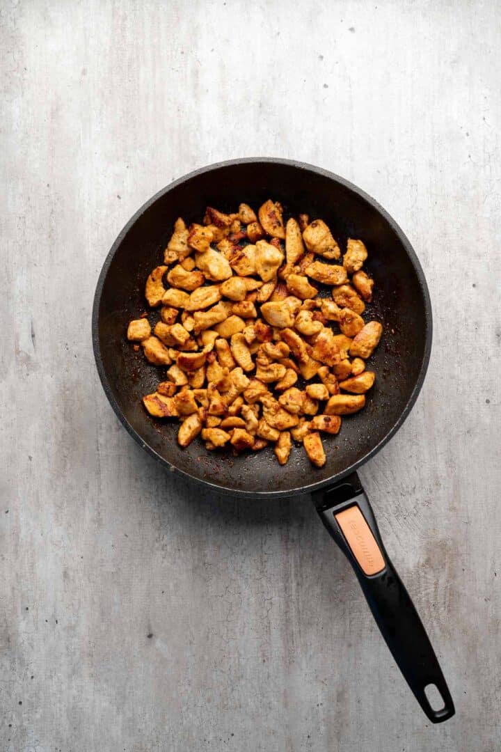Overhead view of cooked, seasoned chicken pieces in a black frying pan on a light countertop, perfect for assembling Coconut Chicken Rice Bowls.