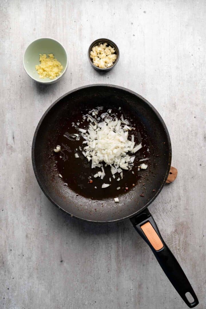 A frying pan with chopped onions inside, next to bowls of minced garlic and chopped ginger on a gray surface—perfect ingredients for Coconut Chicken Rice Bowls.