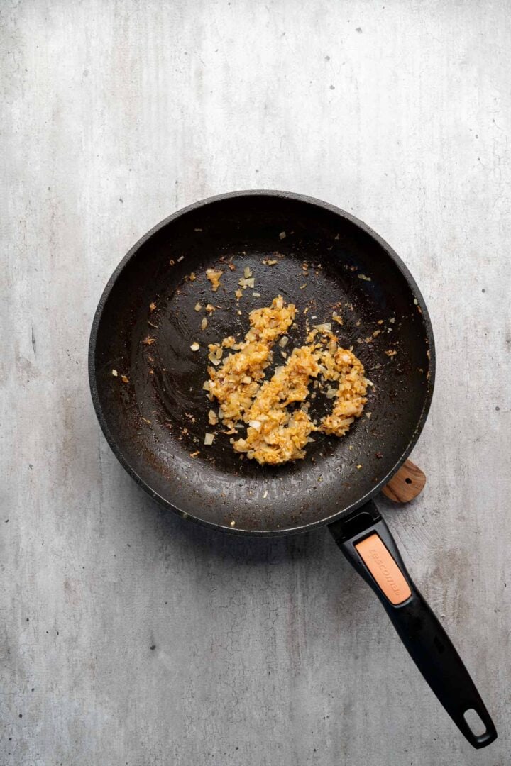 Chopped onions sautéing in a black frying pan on a light gray surface, the perfect start for flavorful Coconut Chicken Rice Bowls.
