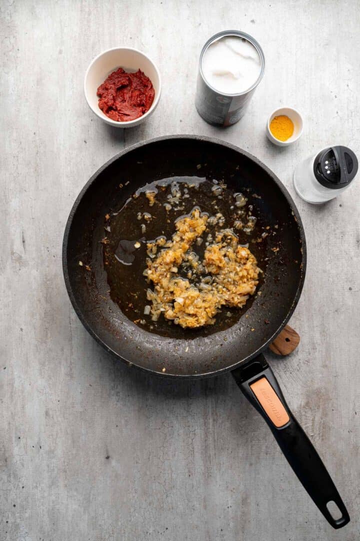A frying pan with sautéed onions; nearby are tomato paste, coconut milk, turmeric, and a salt shaker—perfect ingredients for Coconut Chicken Rice Bowls.