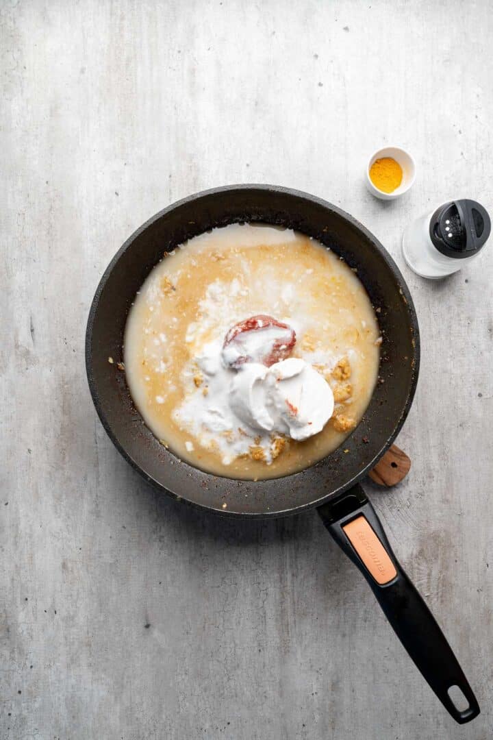 A frying pan with coconut milk, tomato paste, and spices—perfect for making Coconut Chicken Rice Bowls; turmeric and salt in small bowls beside it.
