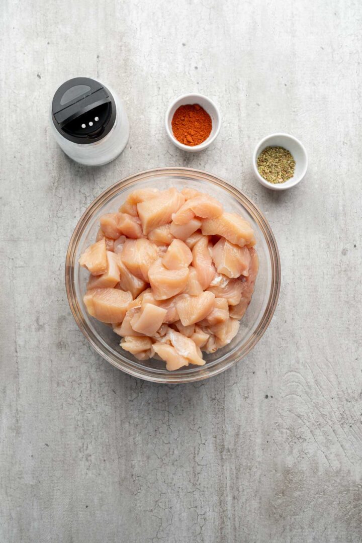 A bowl of raw diced chicken with small bowls of spices and a pepper shaker on a light surface, ready to be transformed into delicious Coconut Chicken Rice Bowls.