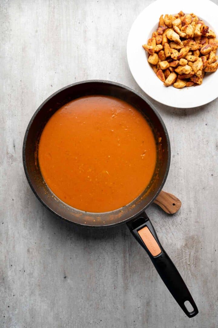 A pan of orange curry sauce sits next to a plate of cooked chicken pieces, perfect for assembling flavorful Coconut Chicken Rice Bowls on a light gray surface.