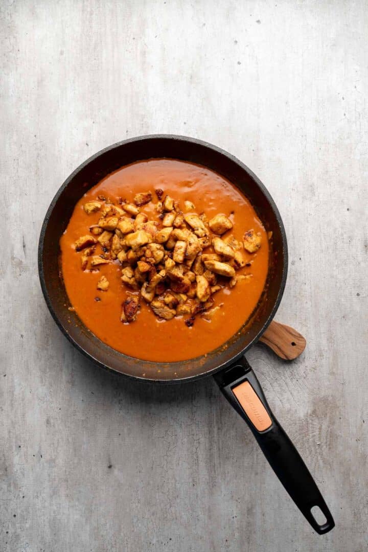 A frying pan with creamy orange sauce and pieces of cooked chicken, perfect for making Coconut Chicken Rice Bowls, sits on a light gray surface.