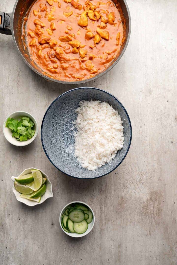 A bowl of white rice beside a pan of curry, with dishes of cilantro, lime wedges, and cucumber slices nearby—perfect fixings for assembling Coconut Chicken Rice Bowls.