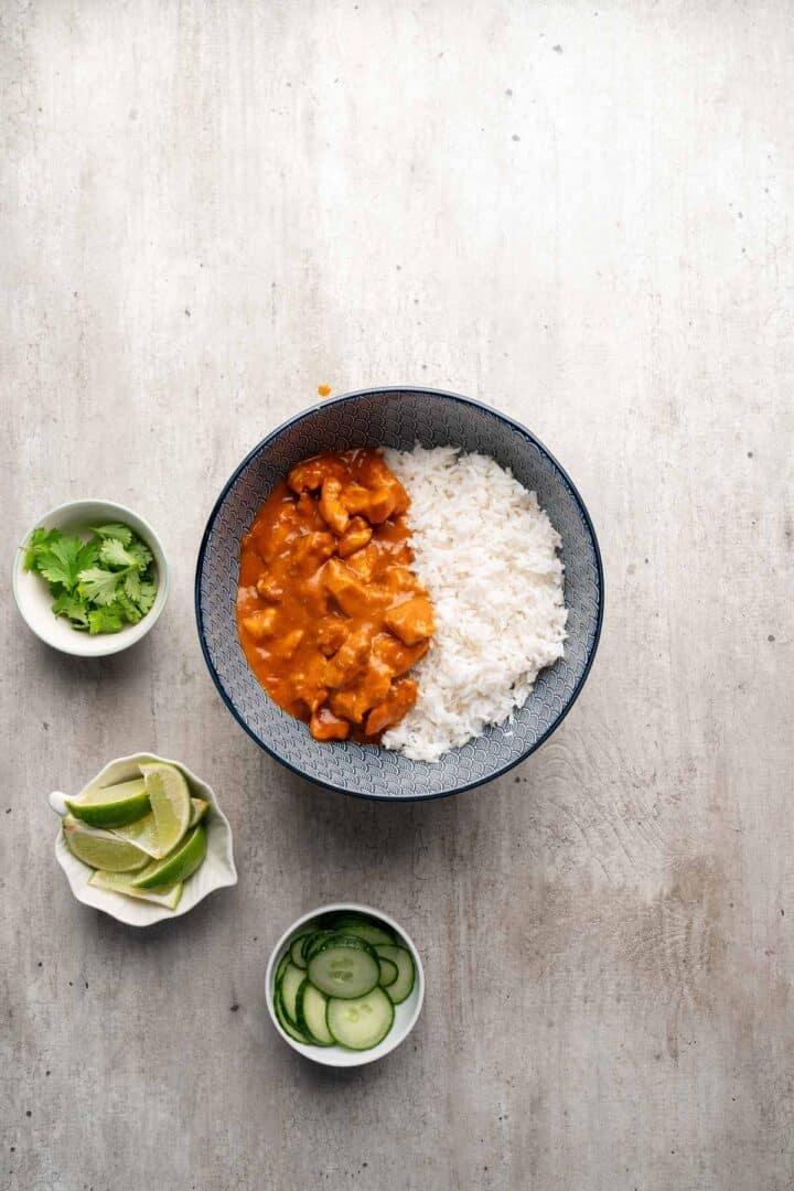 A bowl of rice with orange curry, served with lime wedges, cilantro, and cucumber slices on the side—perfect for creating flavorful Coconut Chicken Rice Bowls.