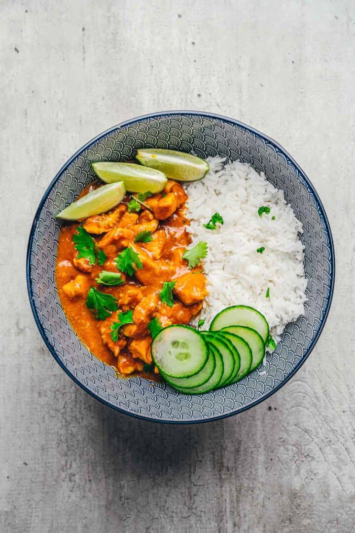 A bowl of Coconut Chicken Rice Bowls with curry, white rice, cucumber slices, lime wedges, and a cilantro garnish.