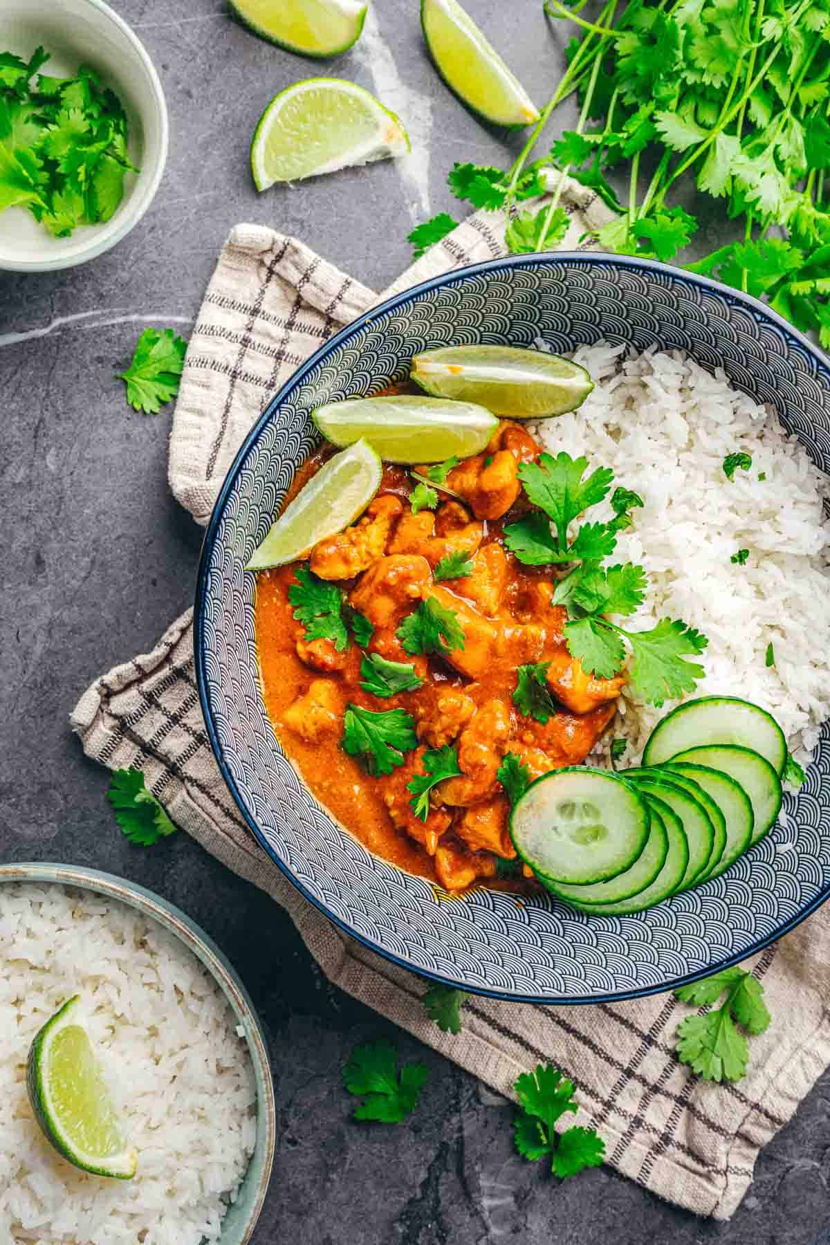 A bowl of Coconut Chicken Rice Bowls with white rice, cucumber slices, lime wedges, and fresh cilantro on a patterned napkin.