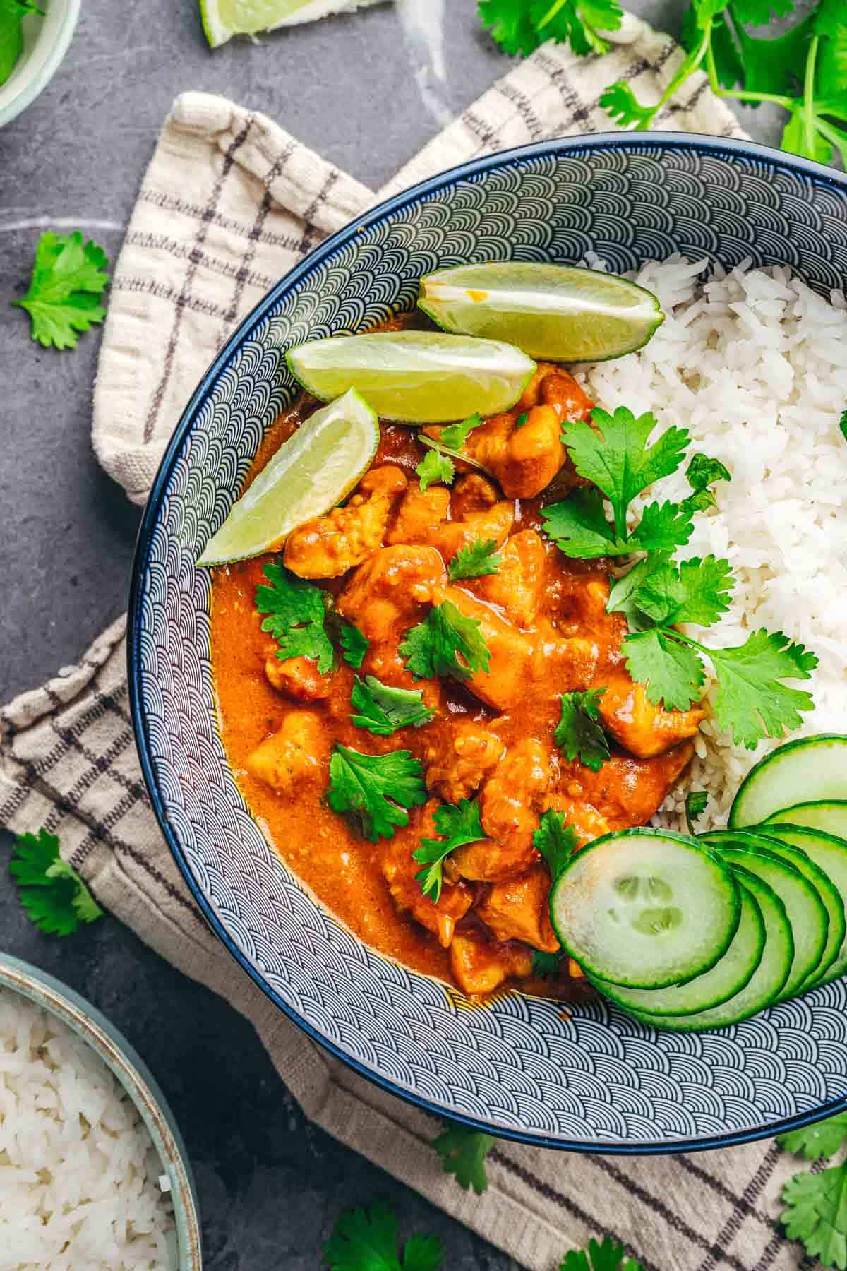 Bowl of Coconut Chicken Rice Bowls with curry chicken, garnished with cilantro, cucumber slices, and lime wedges.