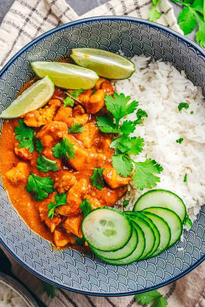 A bowl of Coconut Chicken Rice Bowls with curry chicken, garnished with cilantro, cucumber slices, and lime wedges.