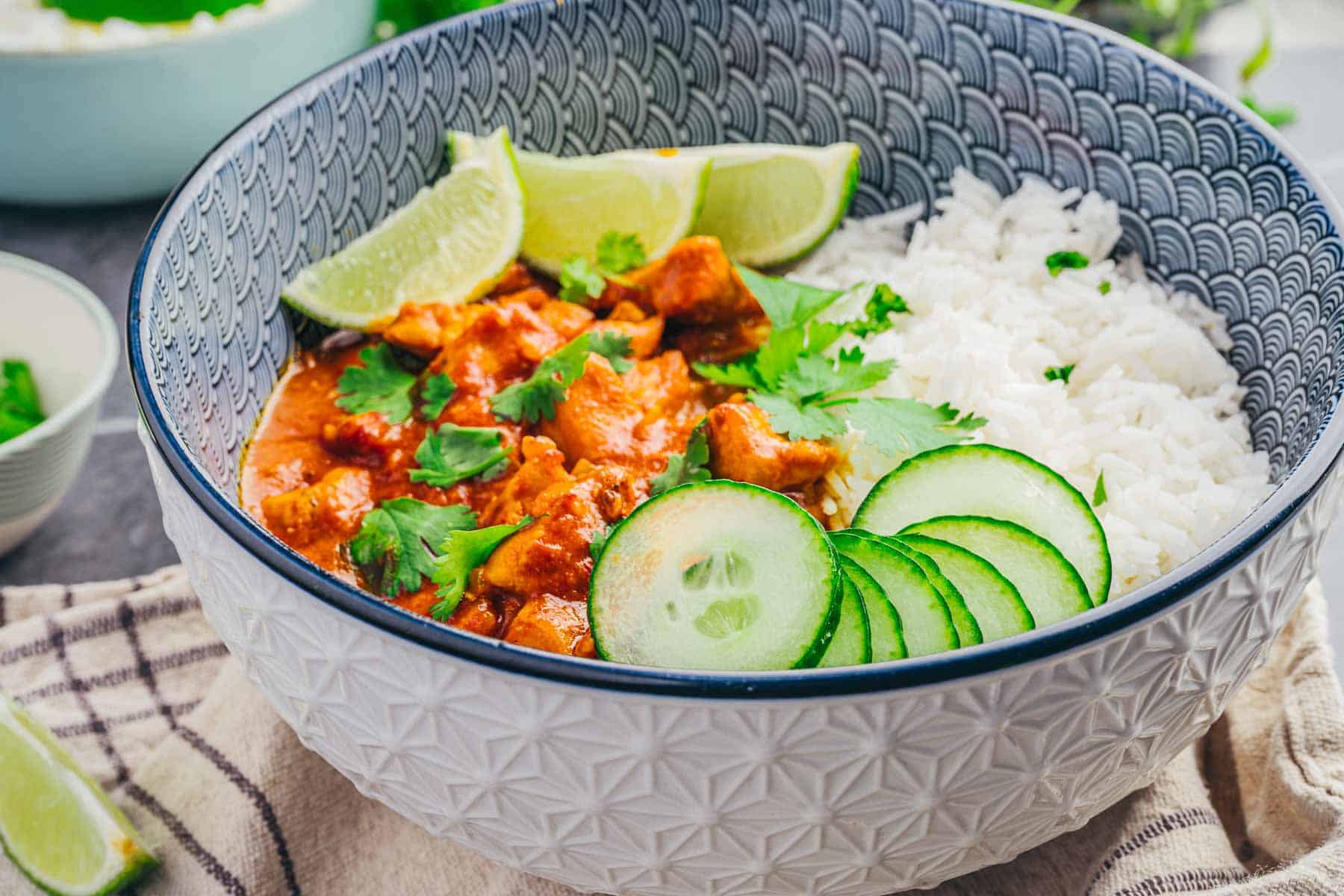 A bowl of Coconut Chicken Rice Bowls with white rice, lime wedges, cilantro, and cucumber slices.
