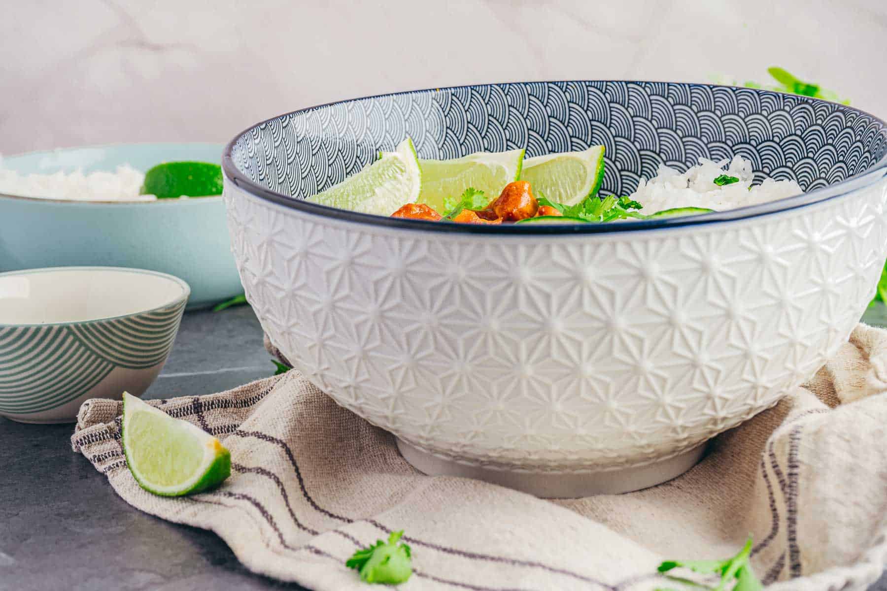 Decorative bowl with Coconut Chicken Rice Bowls, lime wedges, and garnish on a cloth, with more lime and rice bowls in the background.