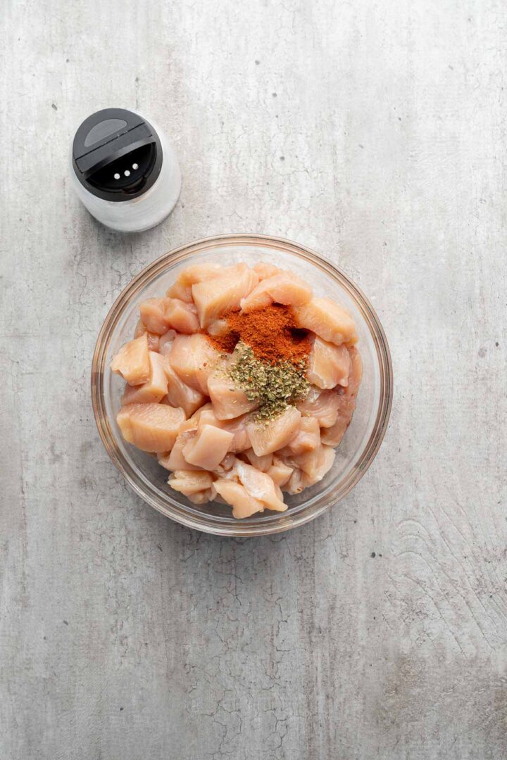 A bowl of diced raw chicken with spices on top, ready to be transformed into delicious Coconut Chicken Rice Bowls, sits next to a salt shaker on a light surface.