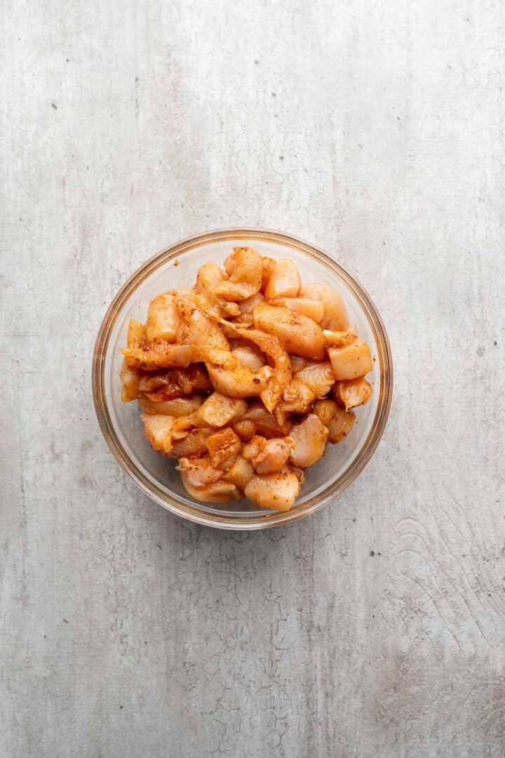 A glass bowl filled with marinated raw chicken pieces, ready to be transformed into delicious Coconut Chicken Rice Bowls, sits on a light gray textured surface.