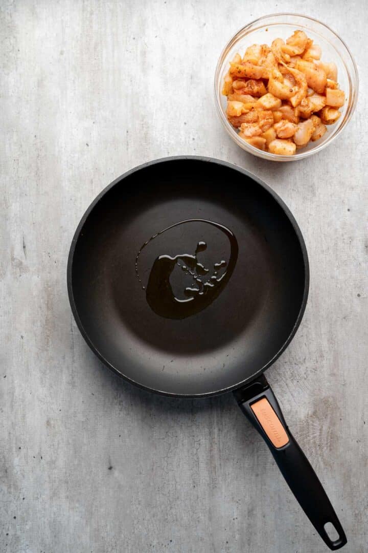 A frying pan with oil and a bowl of seasoned, diced chicken—perfect for starting Coconut Chicken Rice Bowls—sit on a light gray surface.