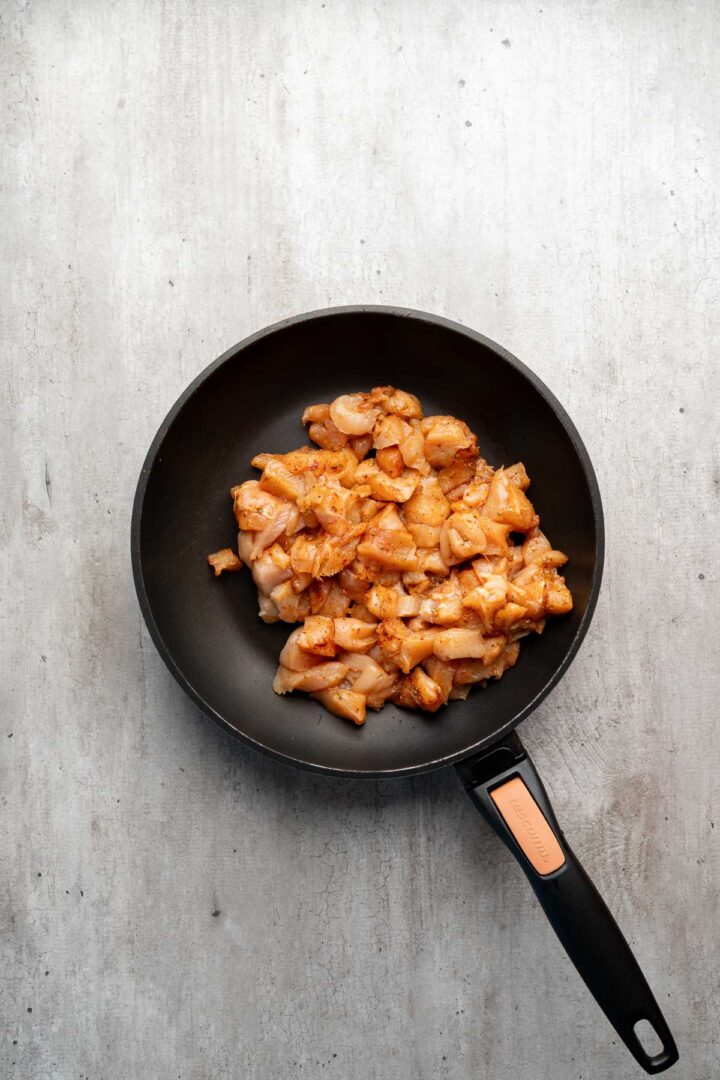 A black frying pan with diced, seasoned raw chicken on a light gray surface, perfect for starting Coconut Chicken Rice Bowls.