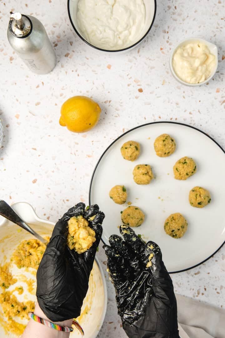 Hands in black gloves shaping falafel mixture into balls over a plate, with bowls and a lemon nearby.