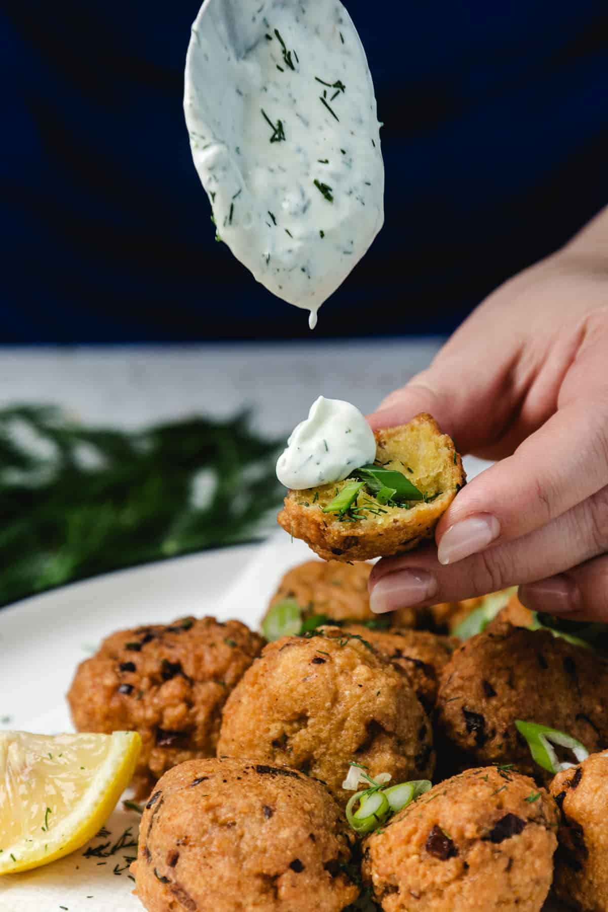 A hand holds a fritter topped with creamy herb sauce, above a plate of fritters and lemon wedges.