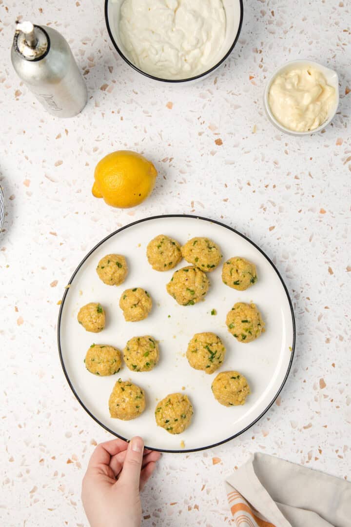 A hand holds a plate of uncooked crab cakes with a lemon and sauces on a white speckled counter.