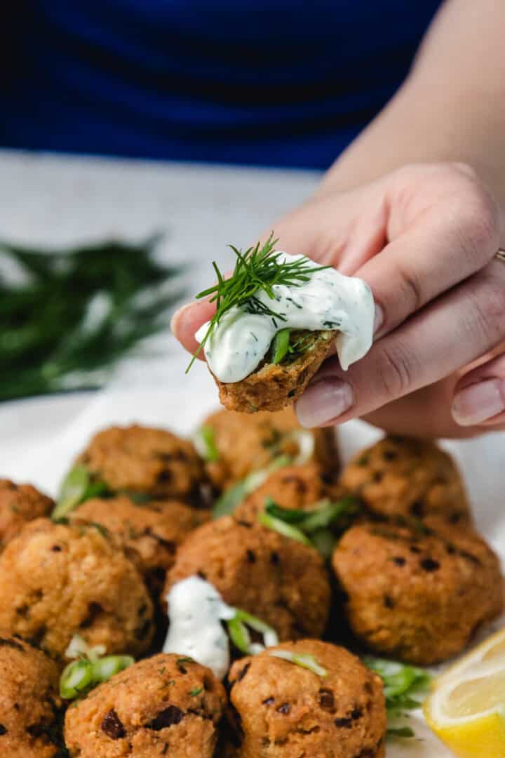 A hand holds a falafel topped with creamy herb sauce over a plate of falafel and lemon slices.