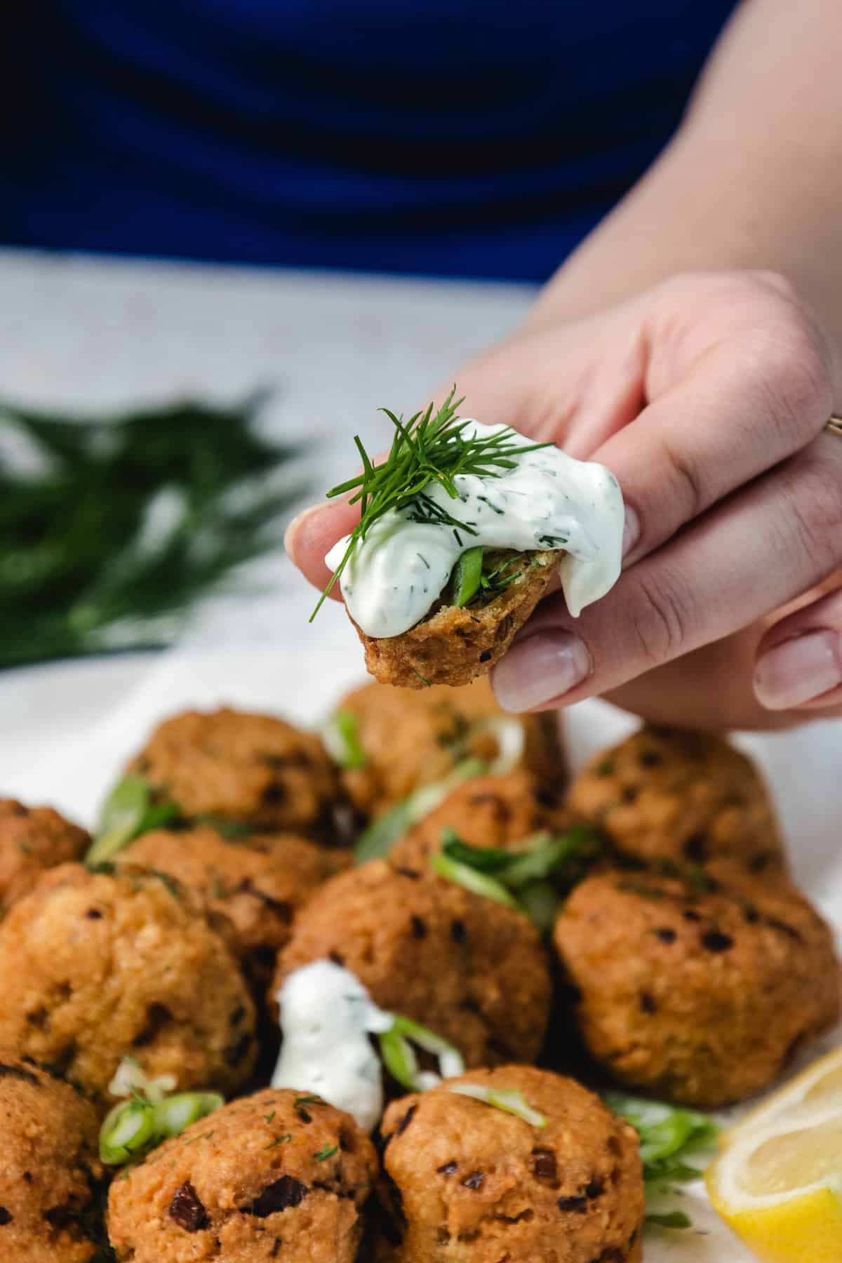 A hand holds a falafel topped with creamy herb sauce over a plate of falafel and lemon slices.