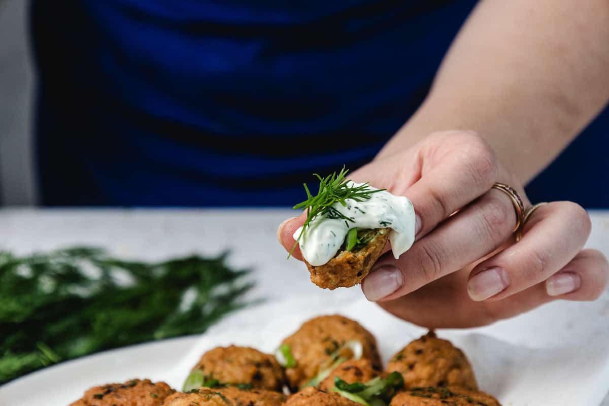 A hand holds a falafel topped with creamy sauce and fresh dill, with more falafel pieces on a plate below.