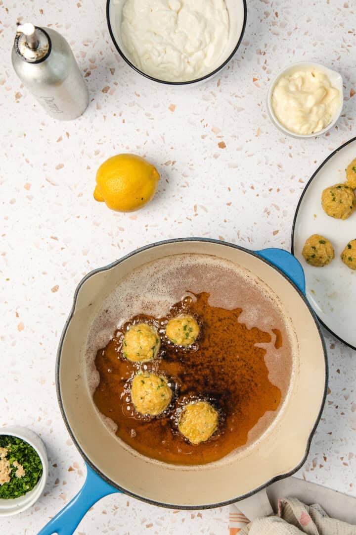Four falafel balls frying in oil in a blue pan, surrounded by bowls of ingredients on a white countertop.