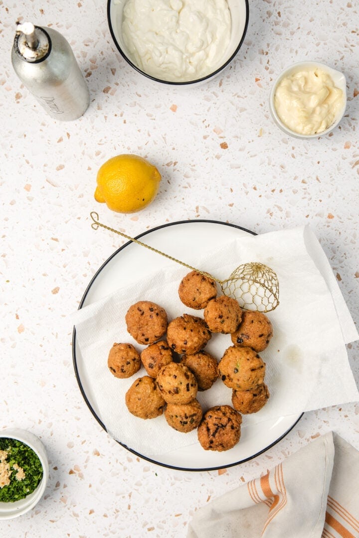 A plate of fried falafel balls on paper towels, surrounded by sauces, a lemon, greens, and a napkin.