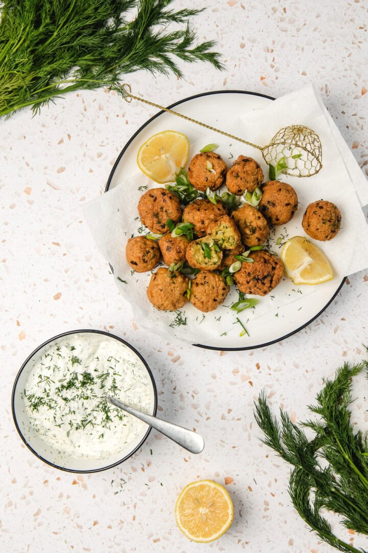 A plate of falafel with lemon wedges and fresh herbs, next to a bowl of creamy dipping sauce with dill.