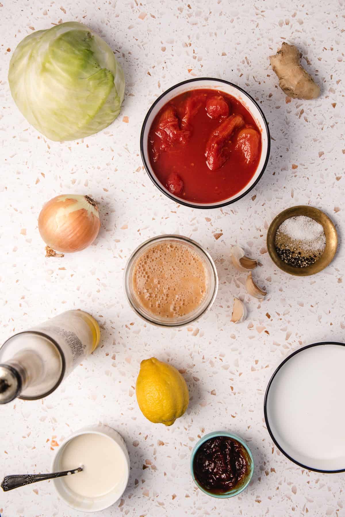 Top-down view of ingredients for Gochujang Tahini Cabbage Soup: cabbage, tomatoes, onion, lemon, garlic, ginger, oil, spices, and sauces arranged on a white surface.