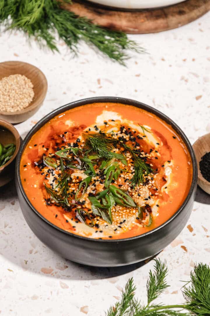 A bowl of creamy tomato soup garnished with seeds, dill, and green onions on a white surface, inspired by the bold flavors of Gochujang Tahini Cabbage Soup.