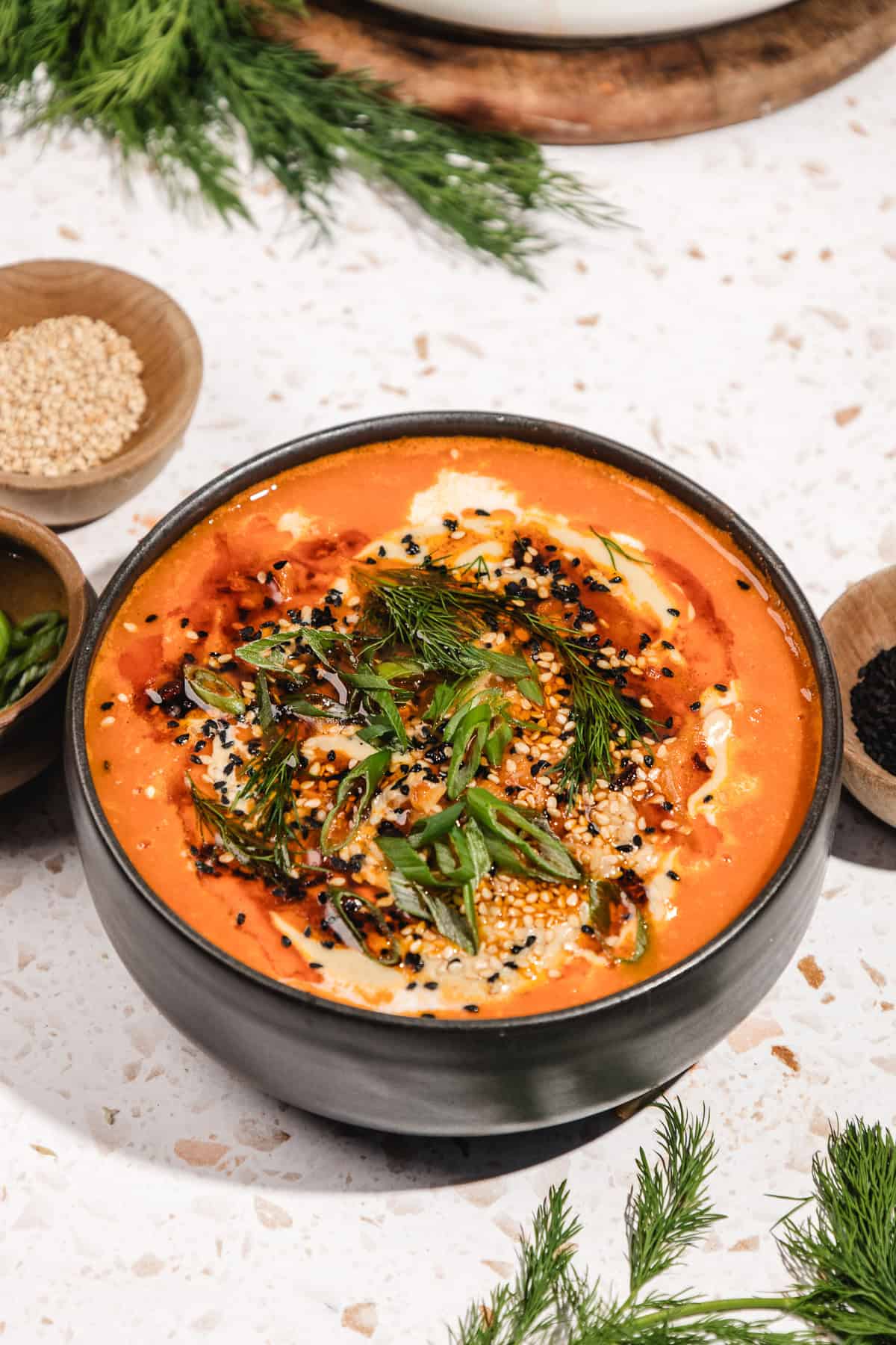 A bowl of creamy tomato soup garnished with seeds, dill, and green onions on a white surface, inspired by the bold flavors of Gochujang Tahini Cabbage Soup.
