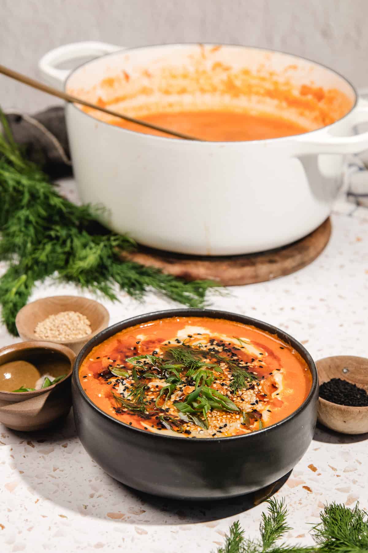 A black bowl of creamy tomato soup topped with herbs and seeds, alongside a pot of Gochujang Tahini Cabbage Soup and fresh dill in the background.