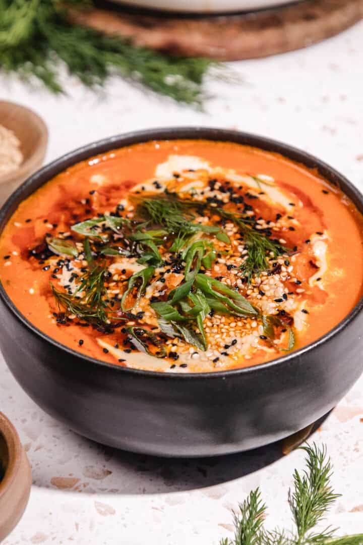 A bowl of creamy Gochujang Tahini Cabbage Soup topped with herbs, seeds, and spices, served in a black bowl on a light surface.