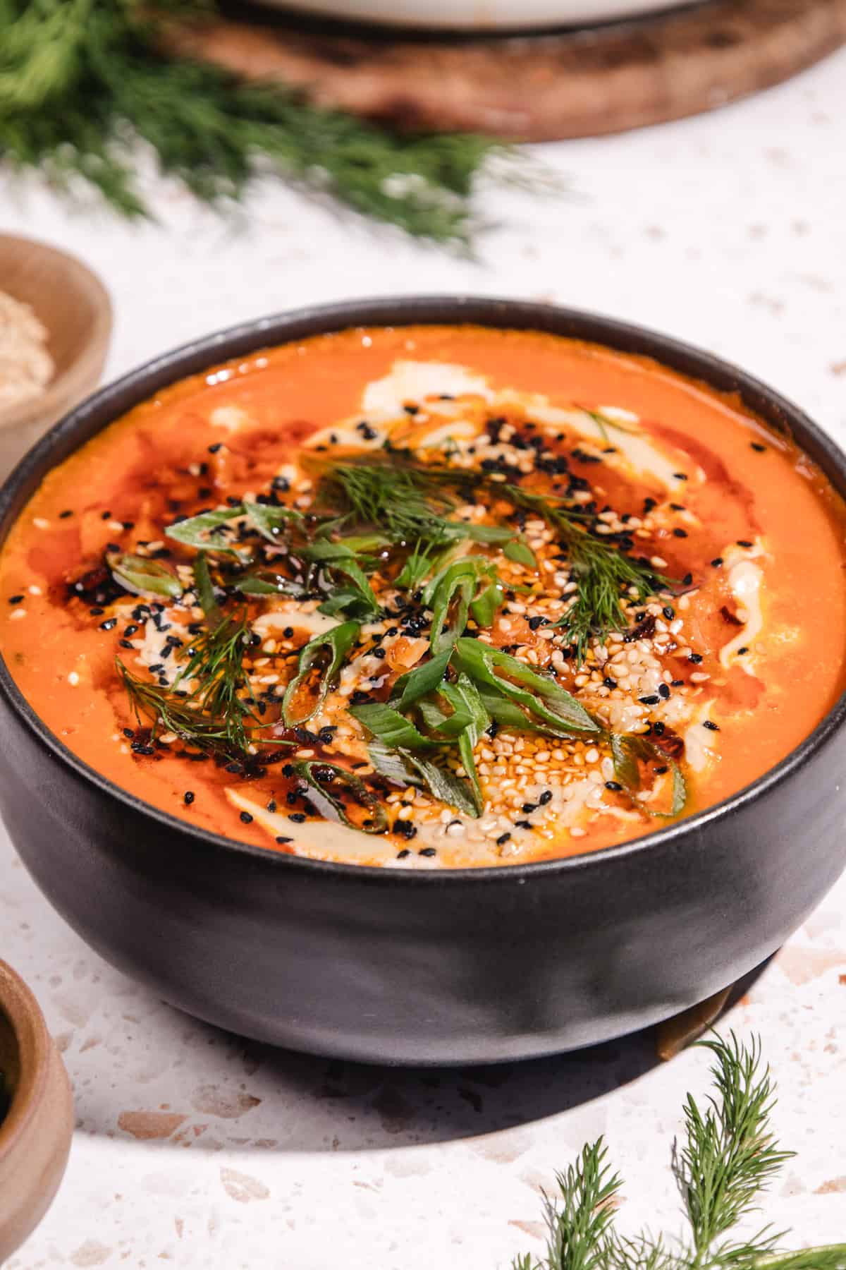 A bowl of creamy Gochujang Tahini Cabbage Soup topped with herbs, seeds, and spices, served in a black bowl on a light surface.