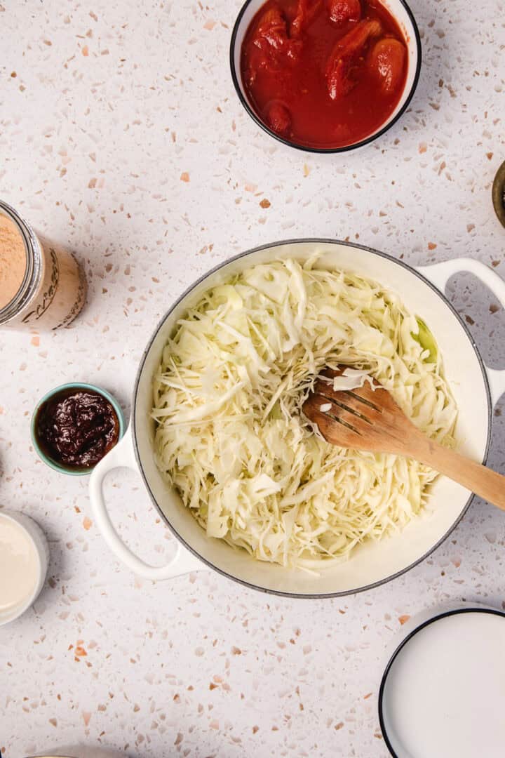 Shredded cabbage in a white pot with a wooden spoon, surrounded by bowls of sauces and ingredients&mdash;perfect for creating a flavorful Gochujang Tahini Cabbage Soup.